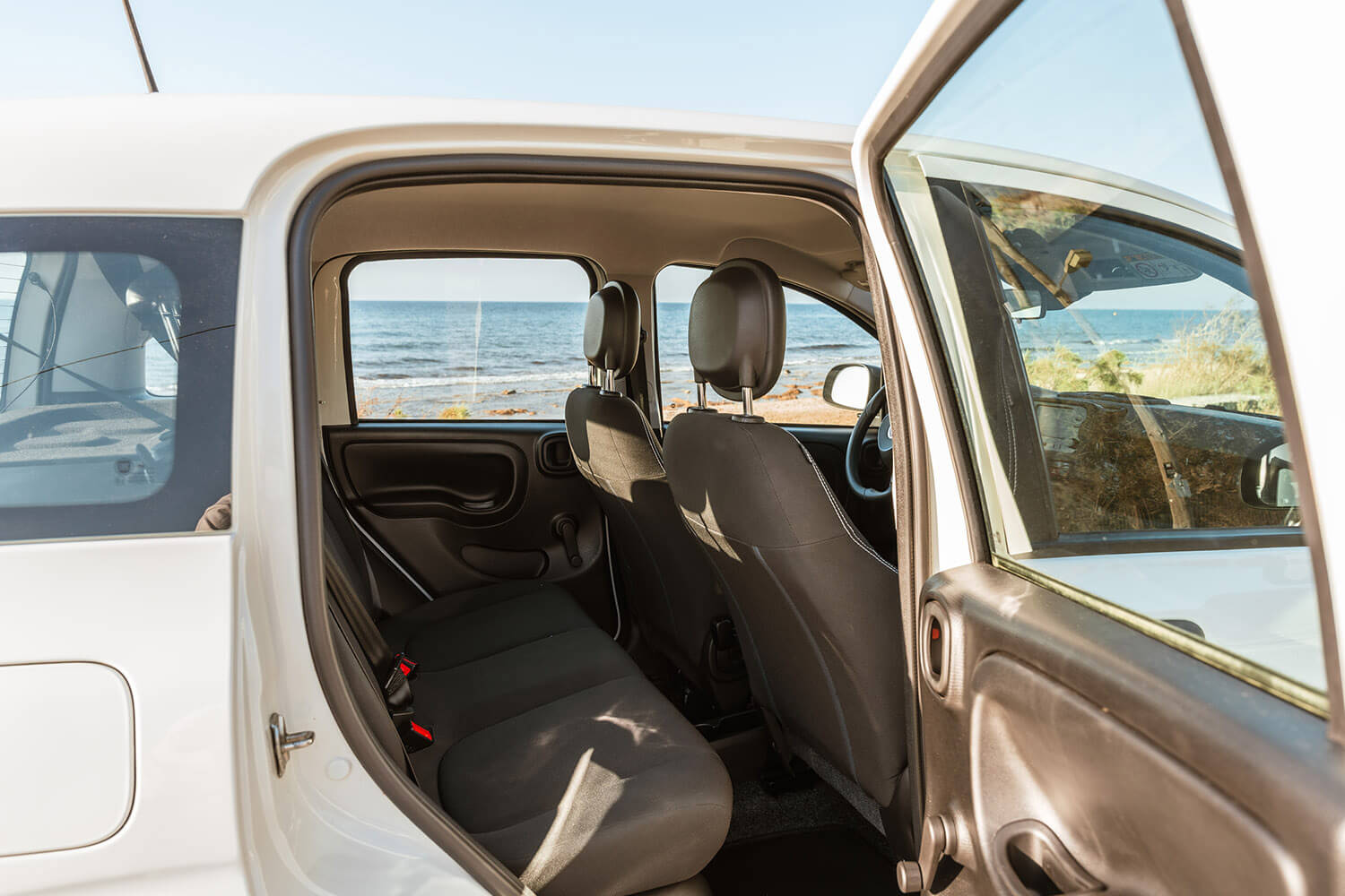 Interior of a white car with black cloth seats; rear door open, beach and sea visible through the windows.