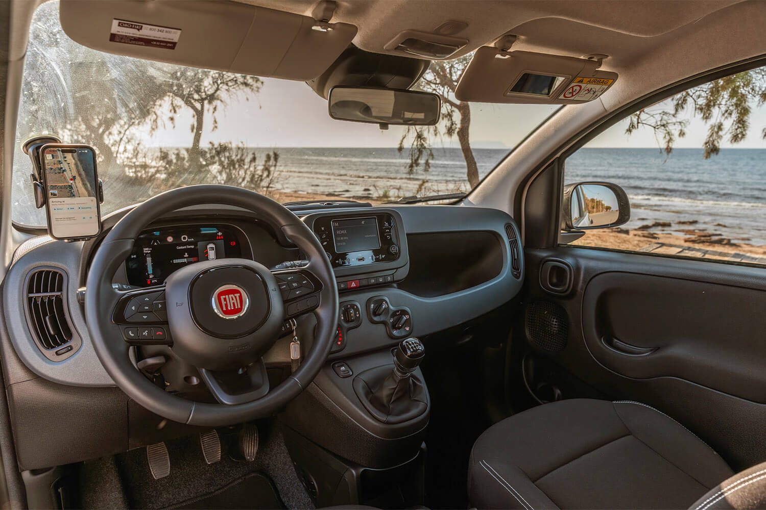 Front interior of a Fiat car by the beach, showing the steering wheel, dashboard, gear stick, and sunlit sea through the windshield on a sunny day.