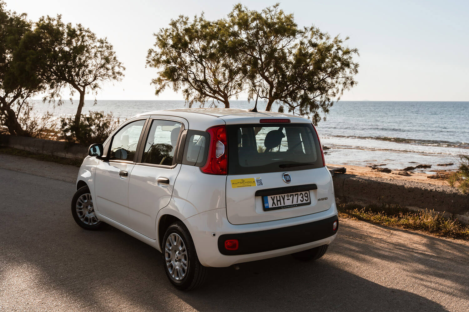White Fiat hatchback parked on a coastal road with the sea and trees in the background.