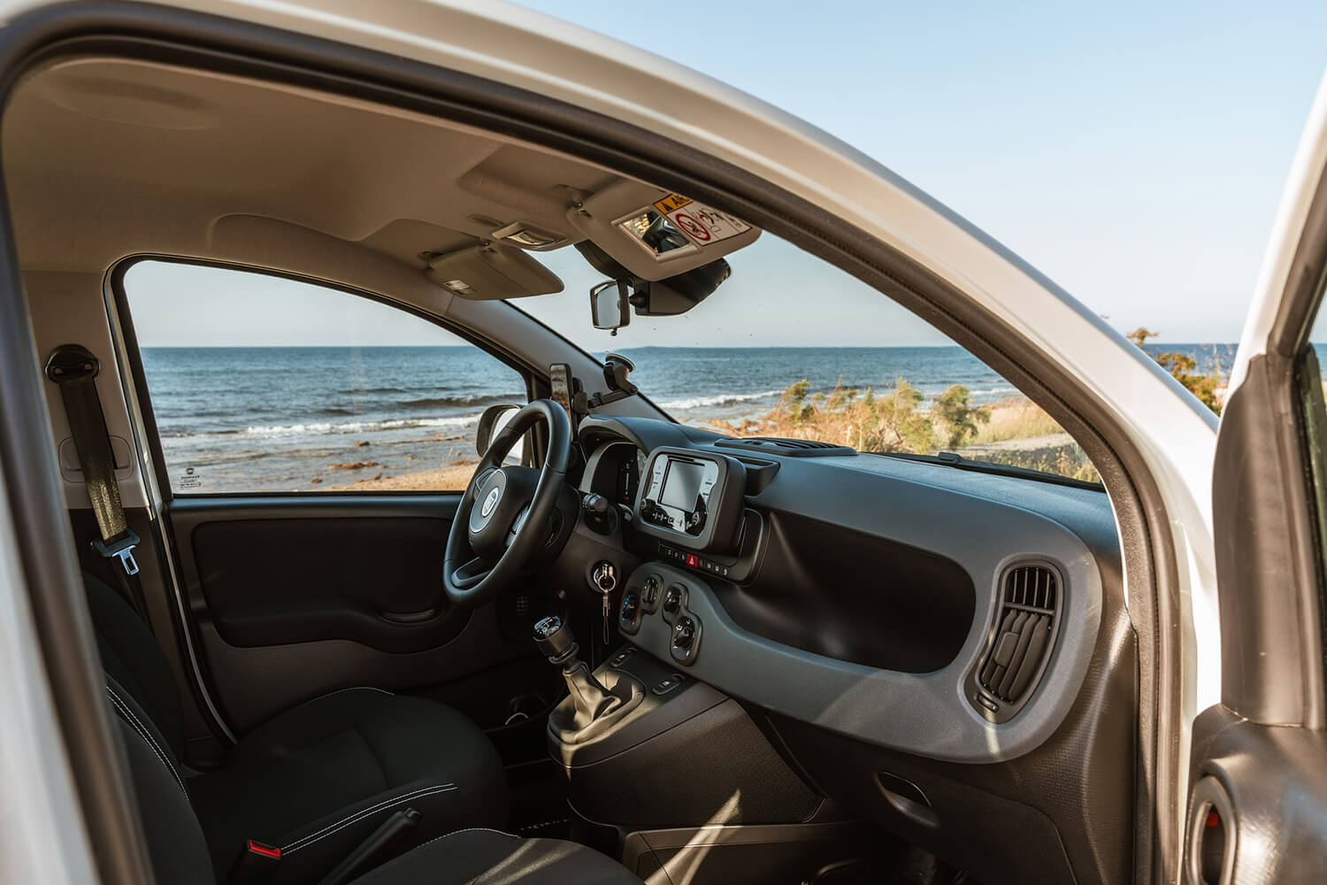 Interior of a car with the steering wheel and dashboard visible, sea and shore seen through the open door window.