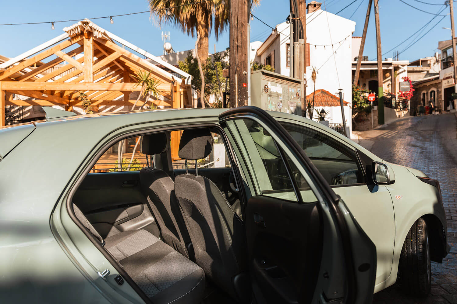Light-green sedan with its passenger door open, showing gray fabric seats, parked on a sunny cobblestone street with a wooden-framed building behind.