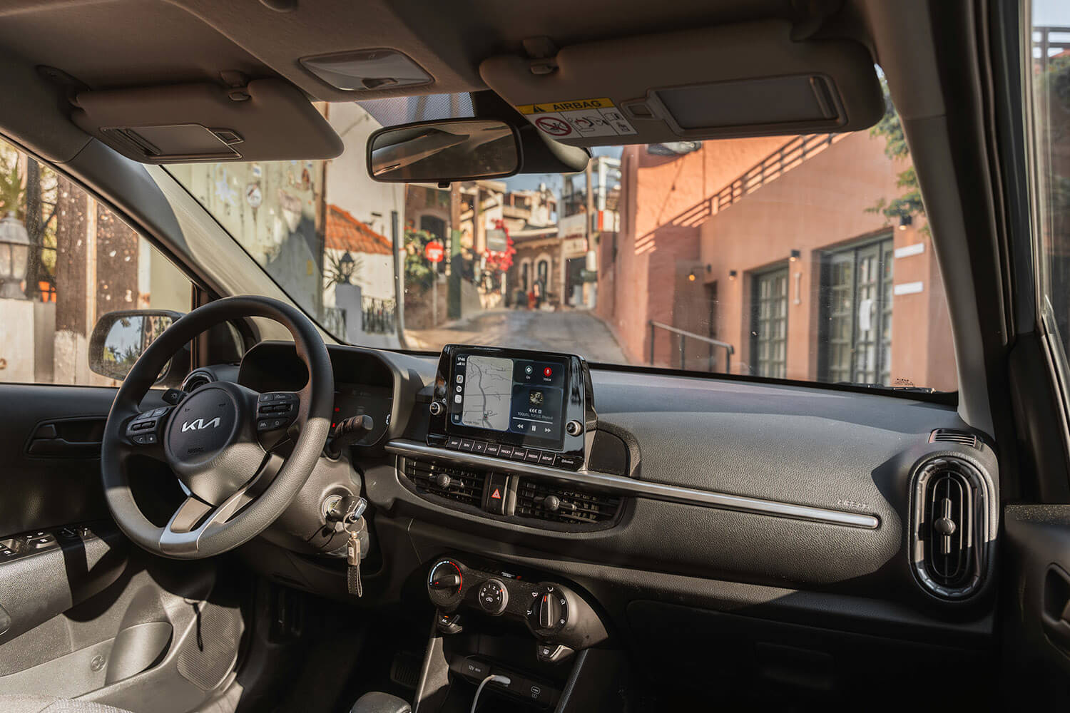 Interior of a car showing steering wheel, center touchscreen, and dashboard; street is visible through the windshield outside the car