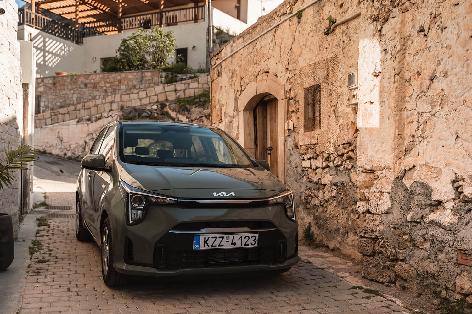 Gray Kia car parked in a narrow cobblestone alley beside old stone walls and a wooden door.