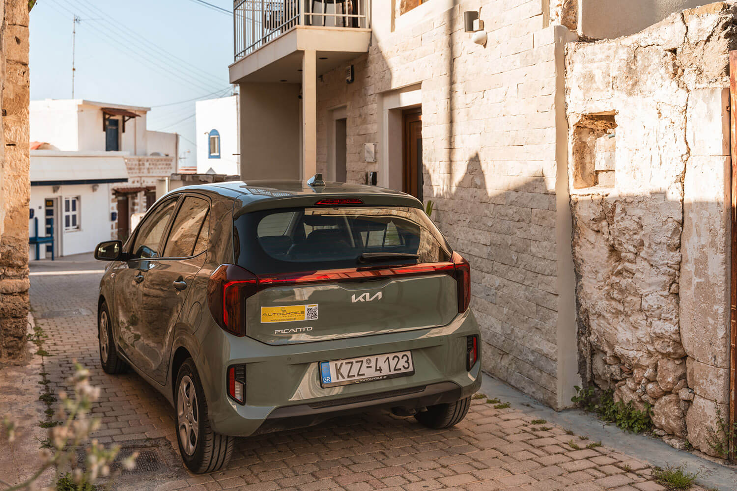 Gray Kia hatchback parked in a narrow stone-walled alley in Greece, rear view with license plate KZZ-4123 and sticker on the back window.