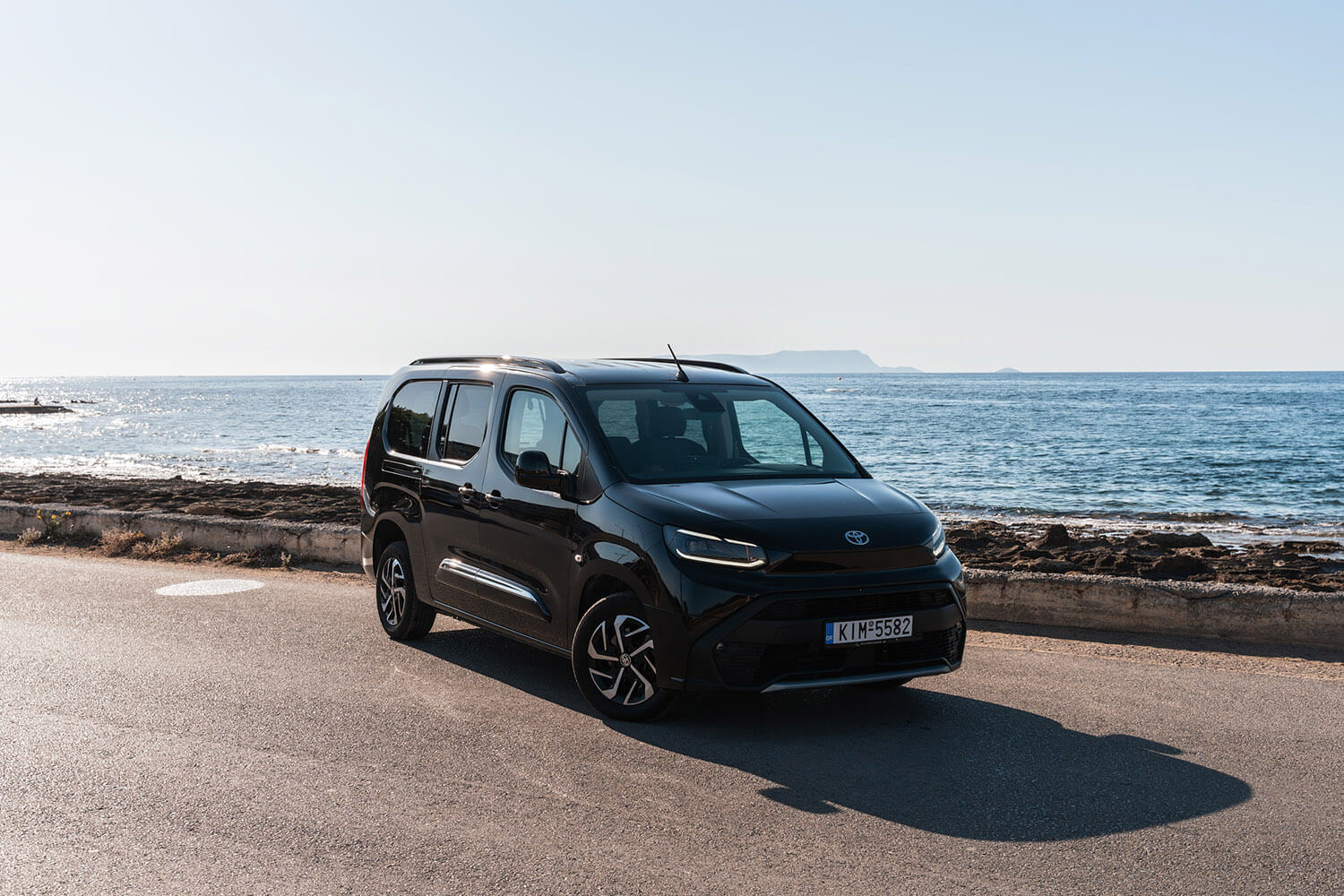 Black Toyota minivan parked on a coastal road with the sea and a distant island in the background, sunny day
