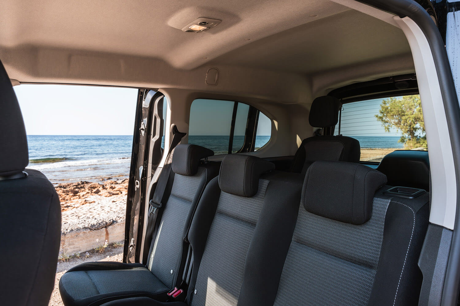 Interior of a car's backseat with gray fabric seats and an open side door, revealing a rocky beach and calm ocean outside.