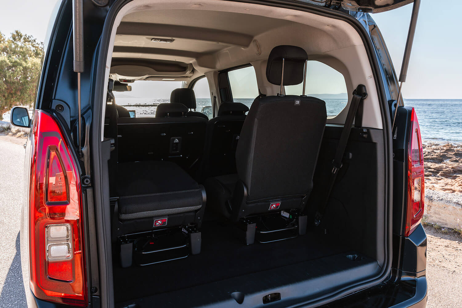 Open rear of a black minivan showing cargo space and two captain chairs, with the sea visible through the back windows.