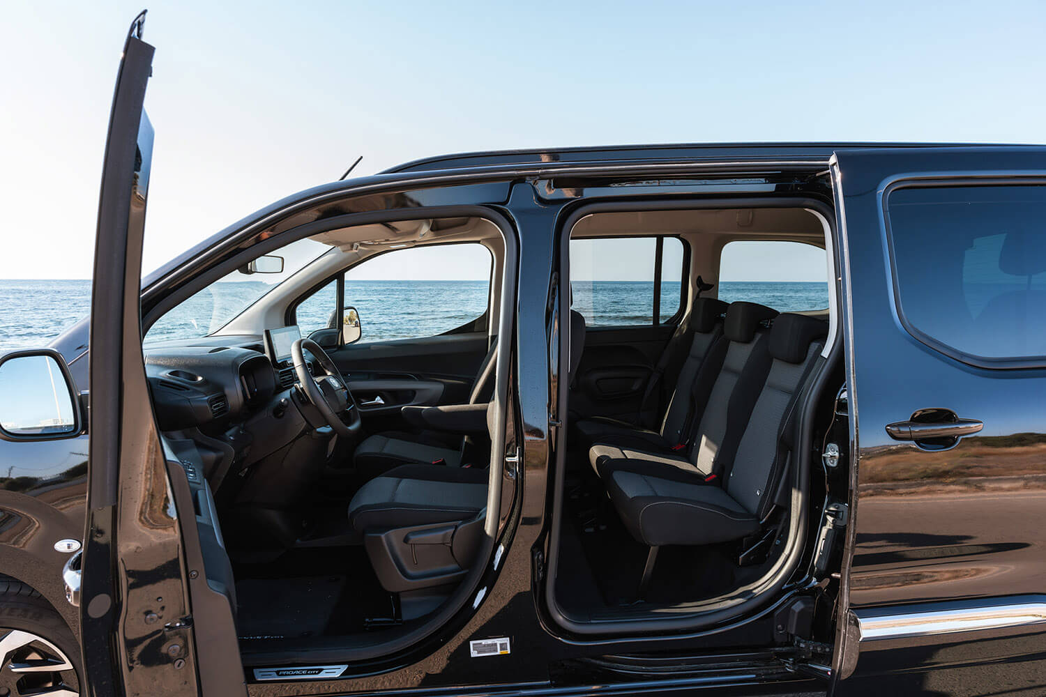 Black passenger van with both side doors open, showing front and second-row seats beside the ocean.