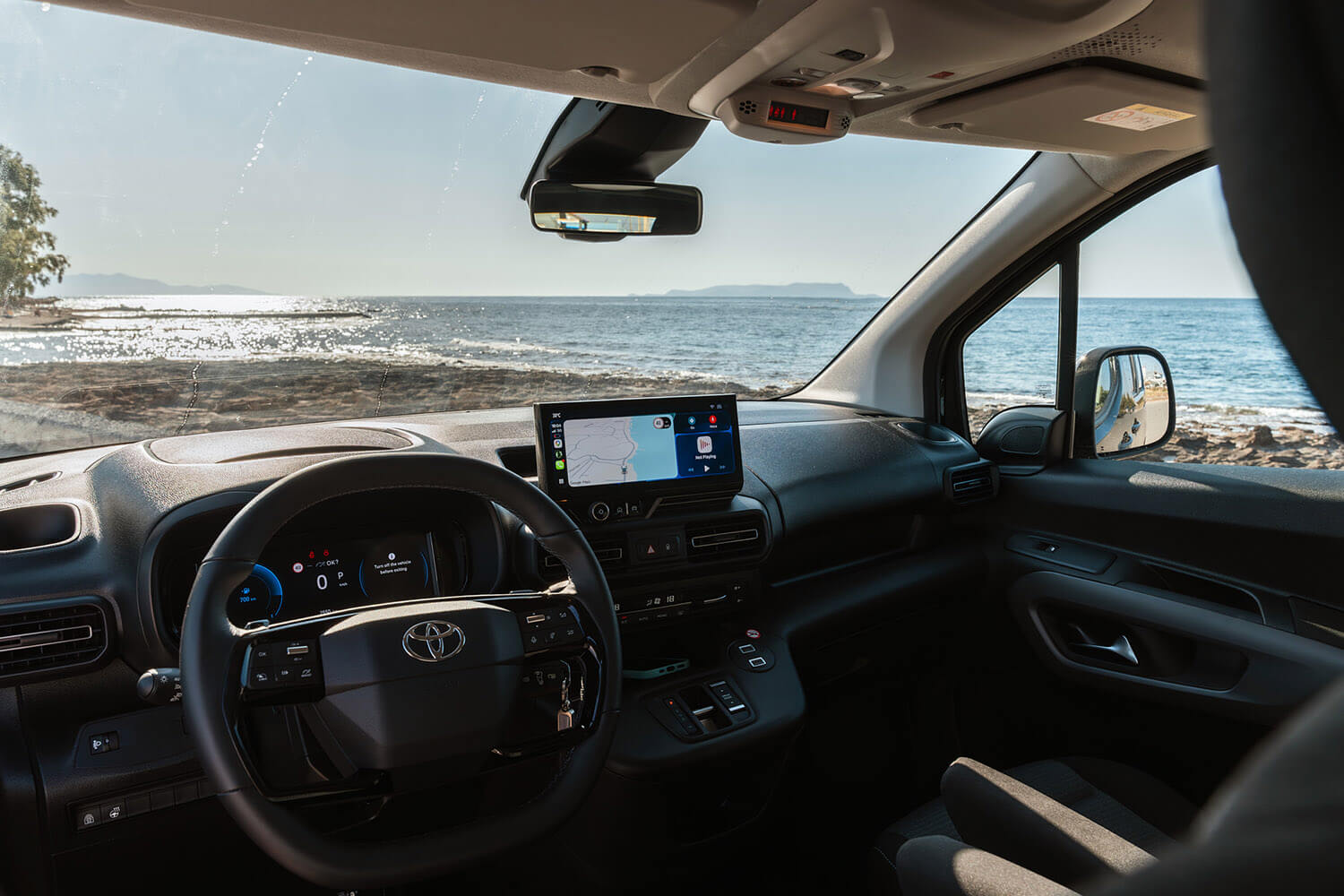 Car interior with steering wheel and center GPS screen, sea view through windshield and coastline ahead.