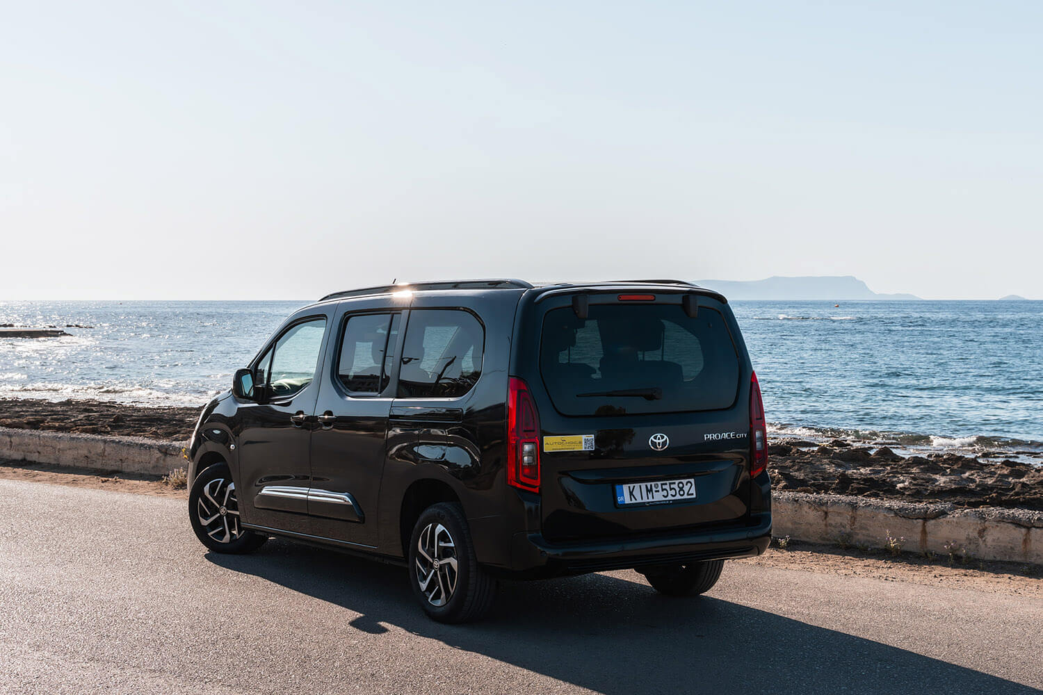 Rear three-quarter view of a black Toyota ProAce City van parked along a coastal road with the sea and distant cliffs in the background
