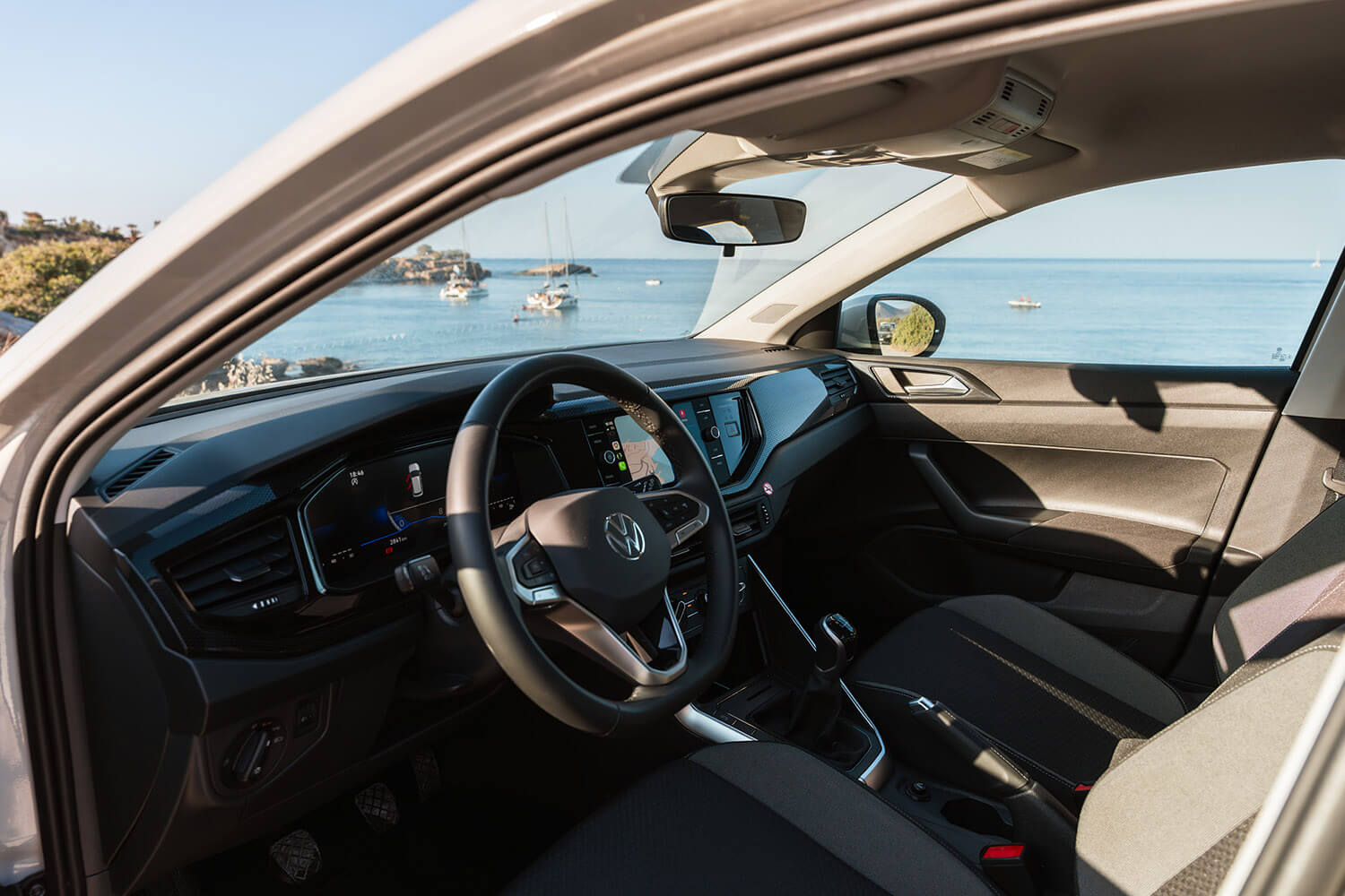 Interior of a modern car with steering wheel, digital dashboard, and view of sea and boats through the open window on a sunny day.