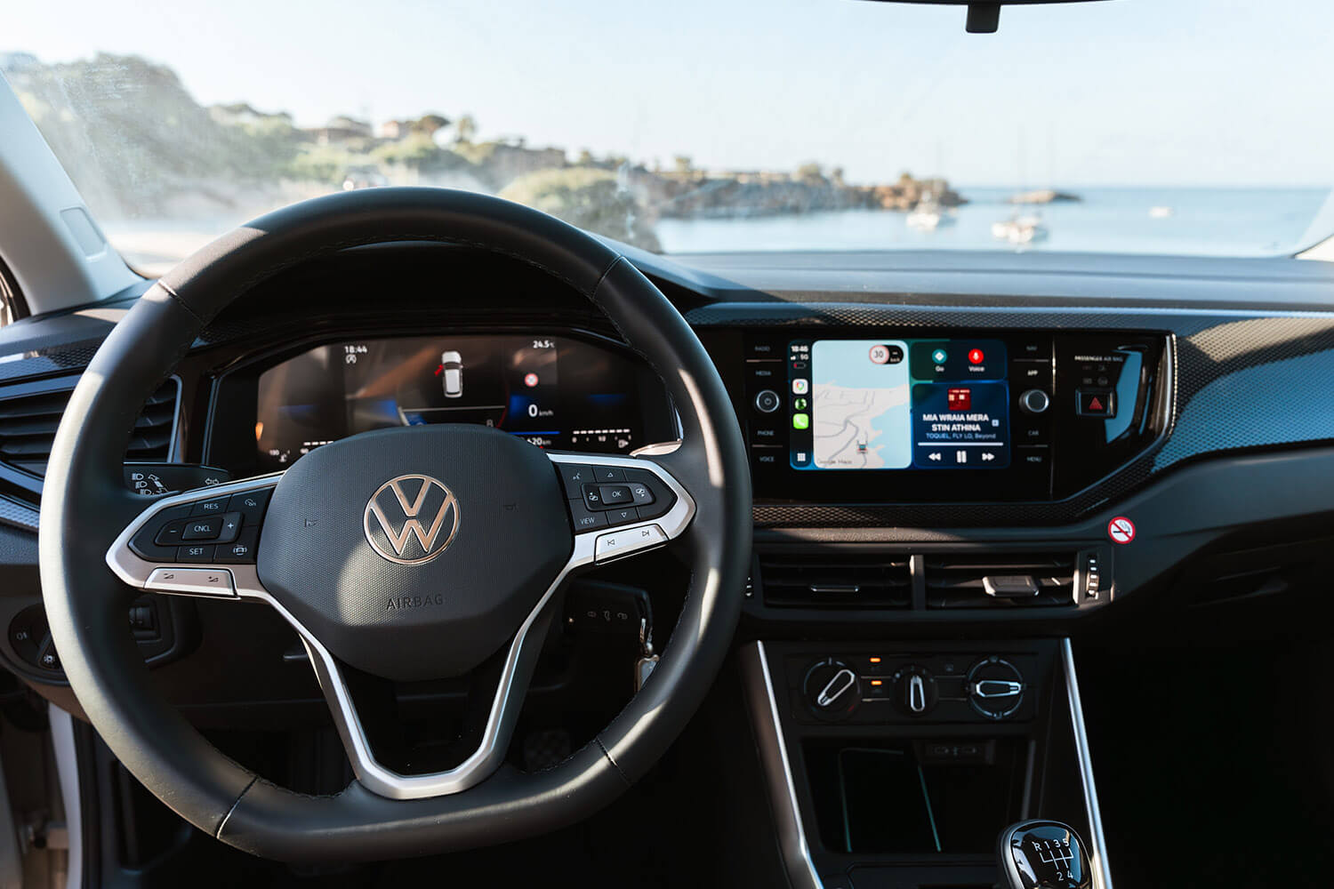VW car cockpit with a digital dashboard and large infotainment screen showing a map; ocean visible through the windshield.
