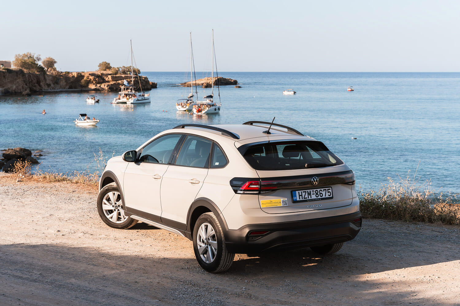 White Volkswagen Taigo parked on a dirt path by the coast with blue sea and sailboats in the background (rear three-quarter view).