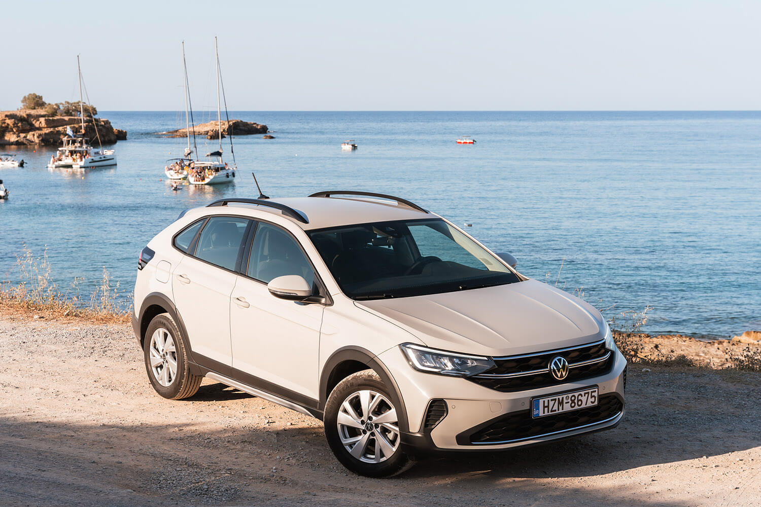 White Volkswagen SUV parked on a rocky coastal road with calm blue sea and sailboats in the background, near the shore.